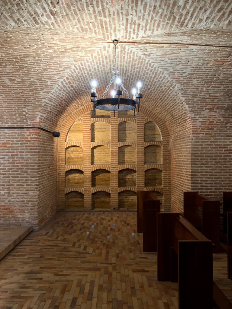 The inside of a Madrid crypt, with burial vaults, pews, arches, and a chandelier.
