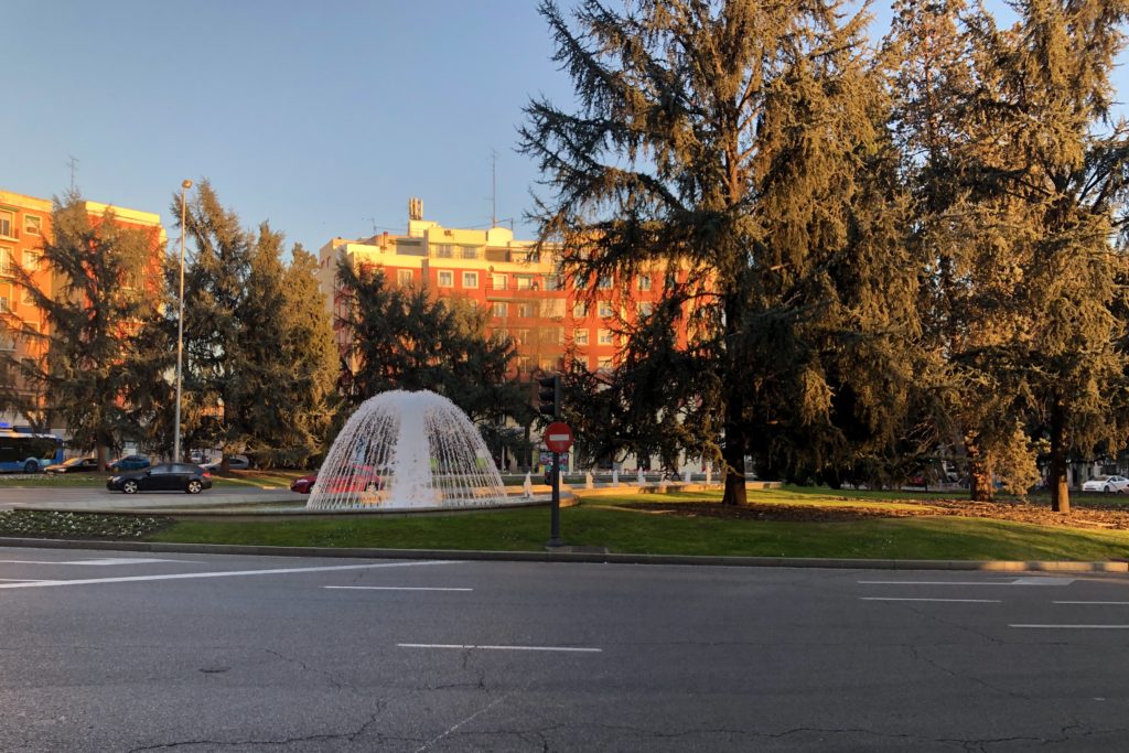 The sun sets on a red building in Madrid, with trees and a fountain in the foreground.