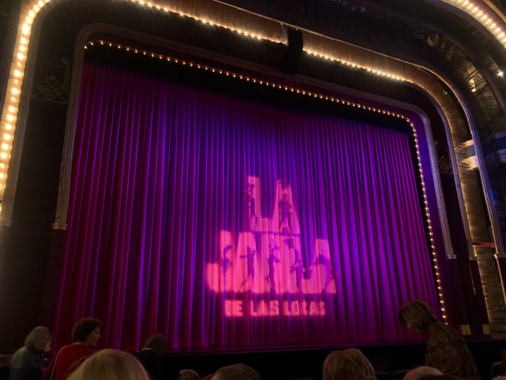 A curtain on a stage reads "La jaula de las locas".
