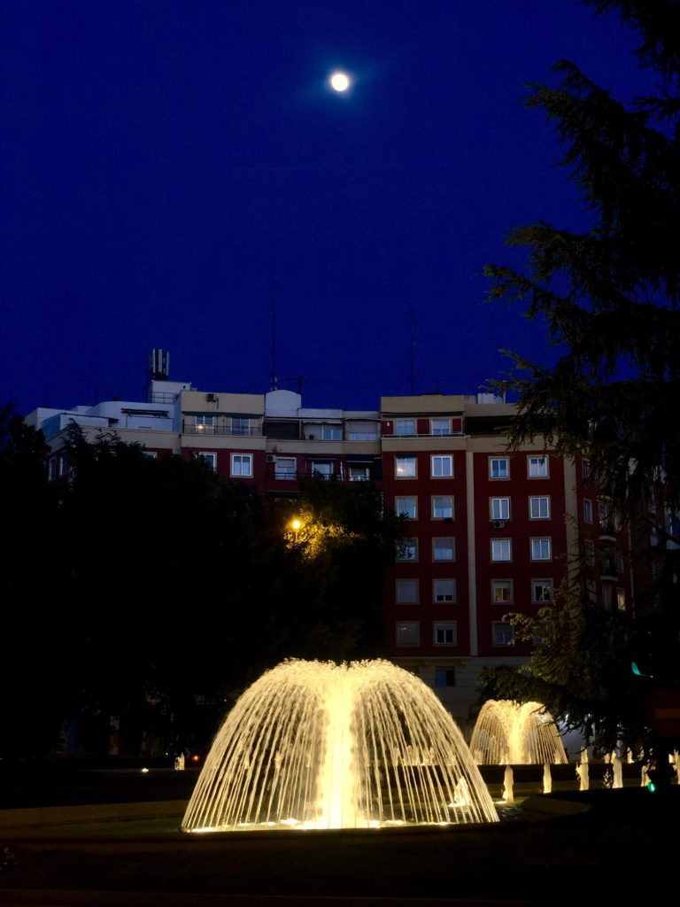 A series of illuminated fountains in Madrid below a full moon sky.