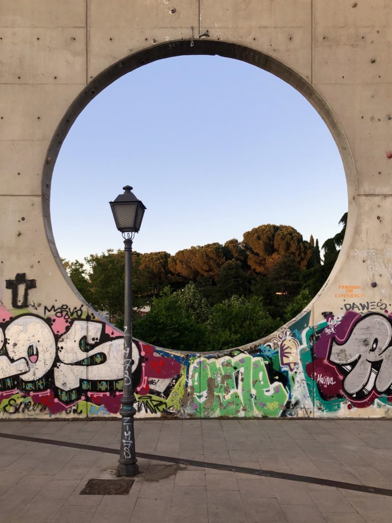 Trees and sky are seen through a circular hole in a concrete wall, with a streetlamp in the foreground.