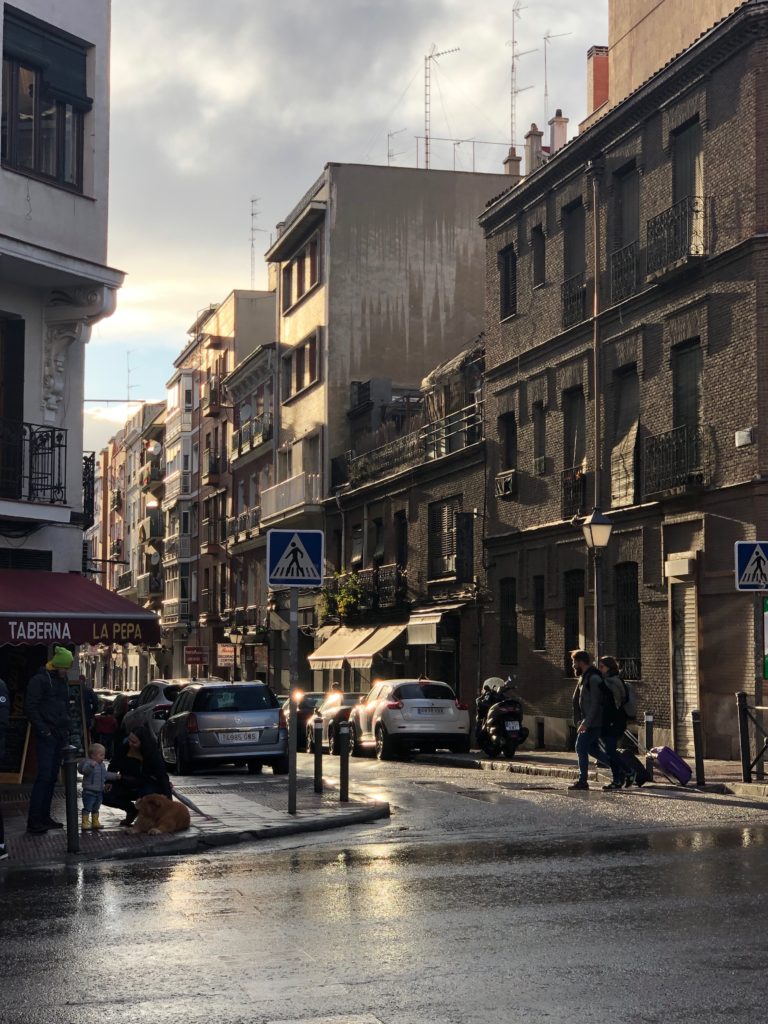 The sun shines down a street, reflecting off the wet road surface and the facades of buildings.