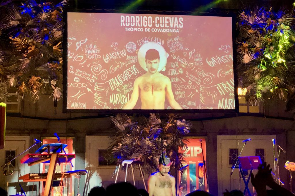 A screen displays Rodrigo Cuevas, with the singer beneath on a stage adorned with flowers.
