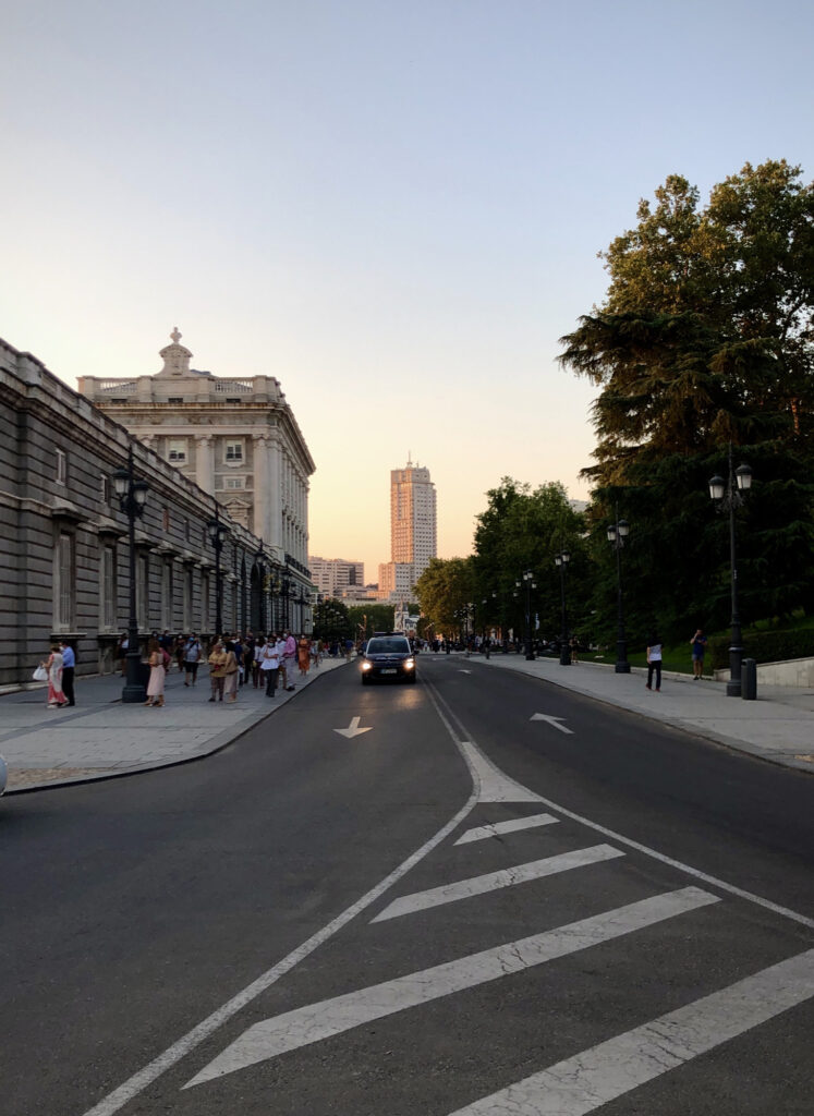 A skyscraper is seen illuminated by the evening sun, with the Royal Palace and a line of trees in the foreground.
